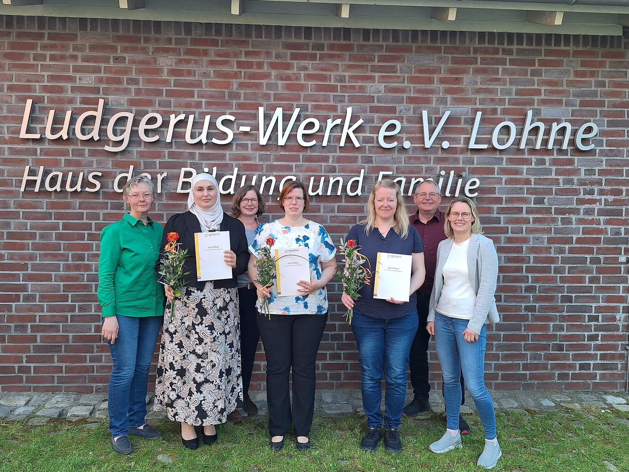 Die Gruppe der Teilnehmerinnen stehen mit den zuständigen Begleitern vor dem Logo an der Außenwand des Ludgerus-Werk. Die Teilnehmerinnen halten ihr Zertifikat und eine rot-orange Rose in den Händen. 