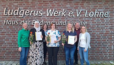 Die Gruppe der Teilnehmerinnen stehen mit den zuständigen Begleitern vor dem Logo an der Außenwand des Ludgerus-Werk. Die Teilnehmerinnen halten ihr Zertifikat und eine rot-orange Rose in den Händen. 