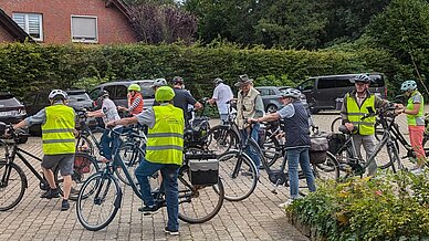 Eine Radfahrgruppe steht auf dem Parkplatz beim Ludgerus-Werk. Die Radfahrer tragen Helme und Warnwesten. 