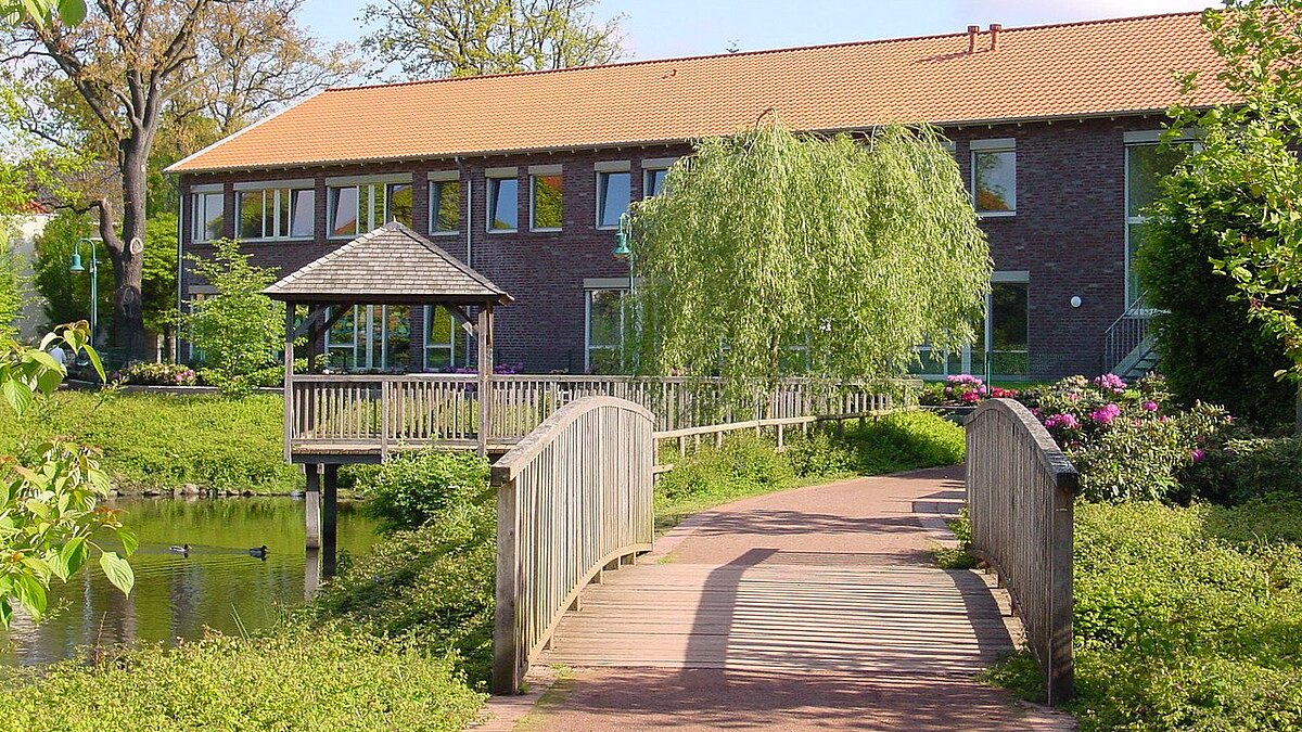 Park hinter dem Ludgeruswerk Man schaut auf den hinteren Teil des Ludgerus-Werk-Gebäudes. Im Vordergrund ist eine kleine Brücke. Links sieht man den Teich und einen Pavillon, der auf das Wasser reicht. Es ist ein sonniger Tag.