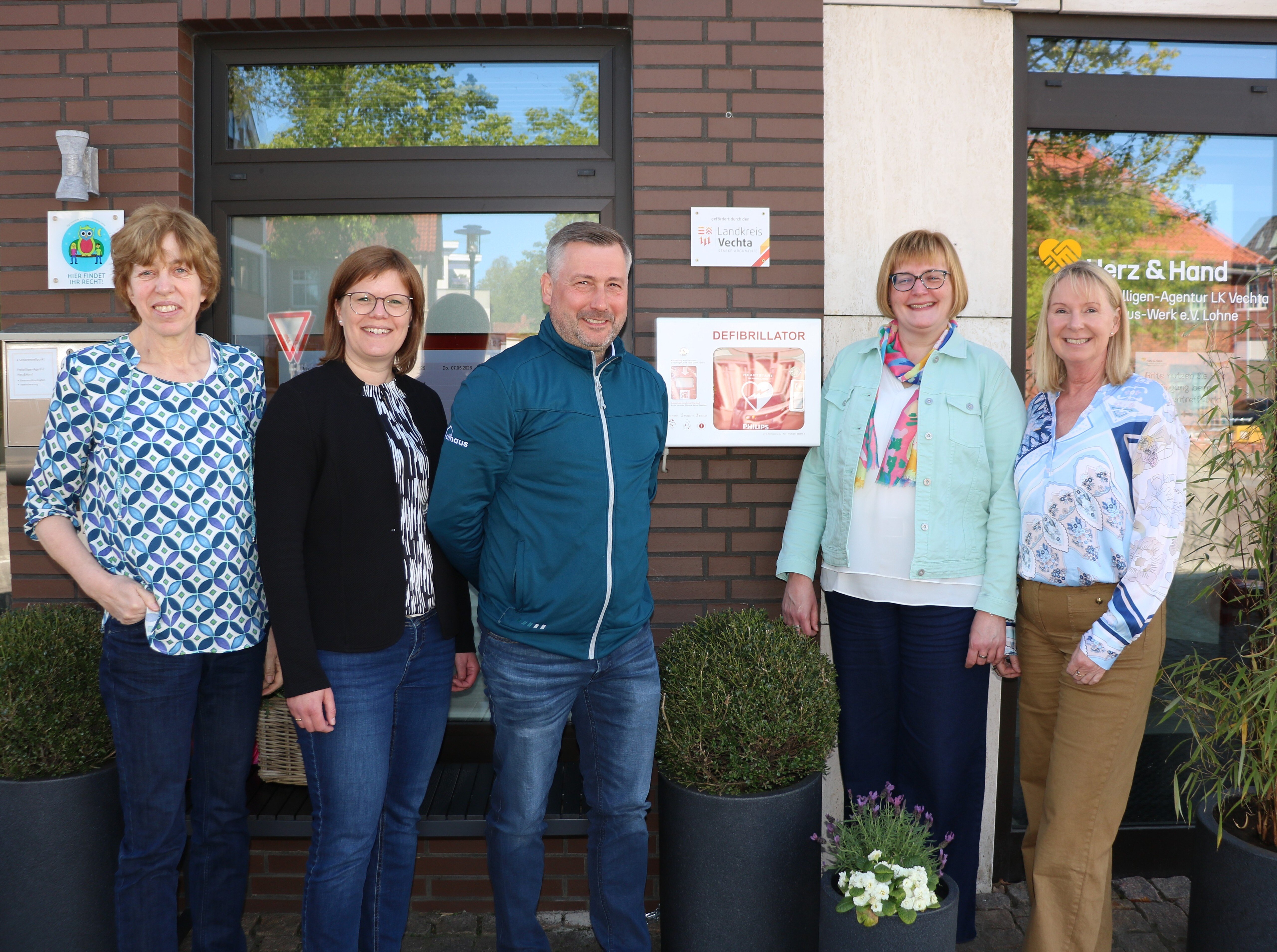 Eine Gruppe Menschen stehen vor einem Außern-Defibrillator und schauen in die Kamera.