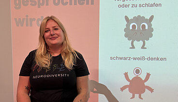 Eine blonde Frau steht vor einer Leinwand. Auf die Leinwand sind Teile ihres Vortrags über Autismus und ADHS projiziert. 