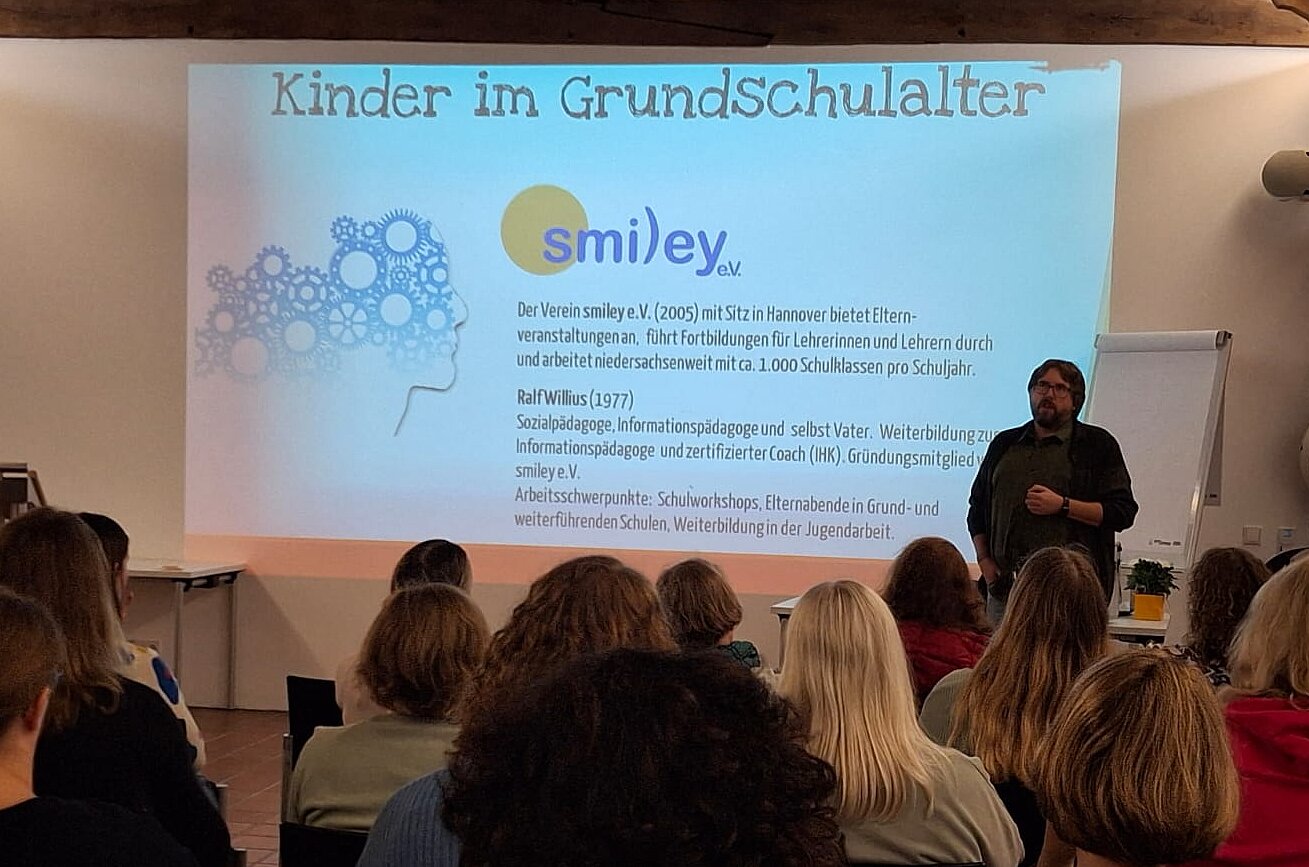 An eine weiße Wand ist eine PowerPointPräsentation projeziert. Daneben steht der Dozent Ralf Wilius. Im Vordergrund sin die Hinterköpfe der Zuhörenden zu sehen. 