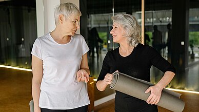 Zwei ältere Damen in Sportbekleidung unterhalten sich in einem Trainingsraum mit Spiegeln. Die rechte Dame hält eine aufgerollte Sportmatte in den Händen.