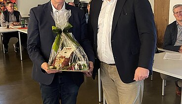 Zwei Männer stehen nebeneinander und lächeln in die Kamera. Der linke hält ein in Folie eingepacktes Geschenk in den Händen, das ihm überreicht worden ist. 