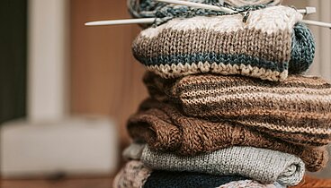Ein Stapel gestrickter Objekte die zusammengelegt auf einem Tisch liegen. Obenauf liegt ein Knäuel Wolle mit Stricknadeln. 