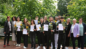 Die Gruppe von Teilnehmenden stehen zusammen mit den Lehrerinnen vor einer Reihe Büsche. Sie halten ihre Abschlusszeugnisse und eine Sonnenblume in den Händen