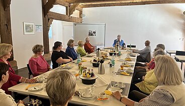 Ein großer Tisch in der Hofstelle im Ludgerus-Werk an dem alle in der Runde sitzen. Es ist für Kaffee und Kuchen gedeckt. An der Stirnseite sitzt die Autorin Maria Meyer, die aus ihrem Buch vorliest.