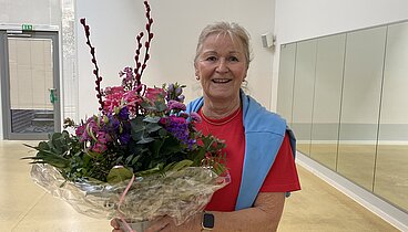 Bild von Doris Trenkamp in der Sporthalle im Ludgerus-Werk. Sie trägt ein rotes Shirt mit einem blauen Pullover über den Schultern. In den Händen hält sie einen großen Blumenstrauß. Die Blumen sind violett-pink und in Klarsichtfolie gepackt. 