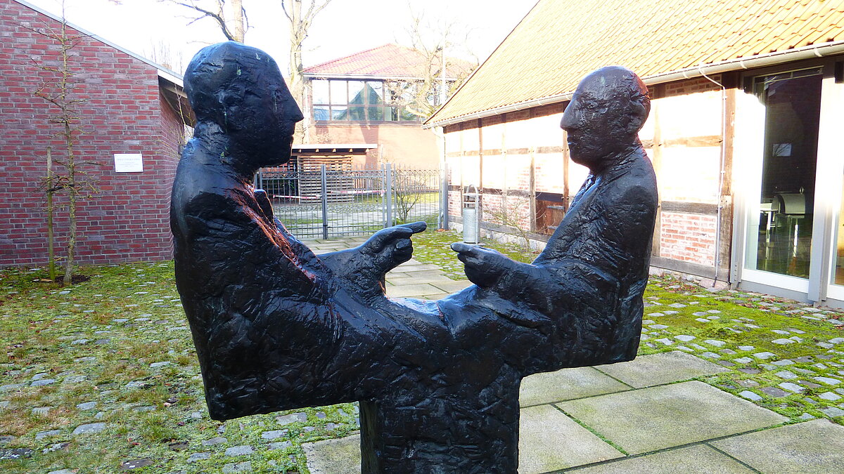 Disput-Skulptur im Innenhof Eine eiserne Skulptur von zwei stilisierten Männern. Sie sehen sich an. Die Männer haben jeweils eine Hand erhoben. Es sieht aus, als ob sie diskutieren.