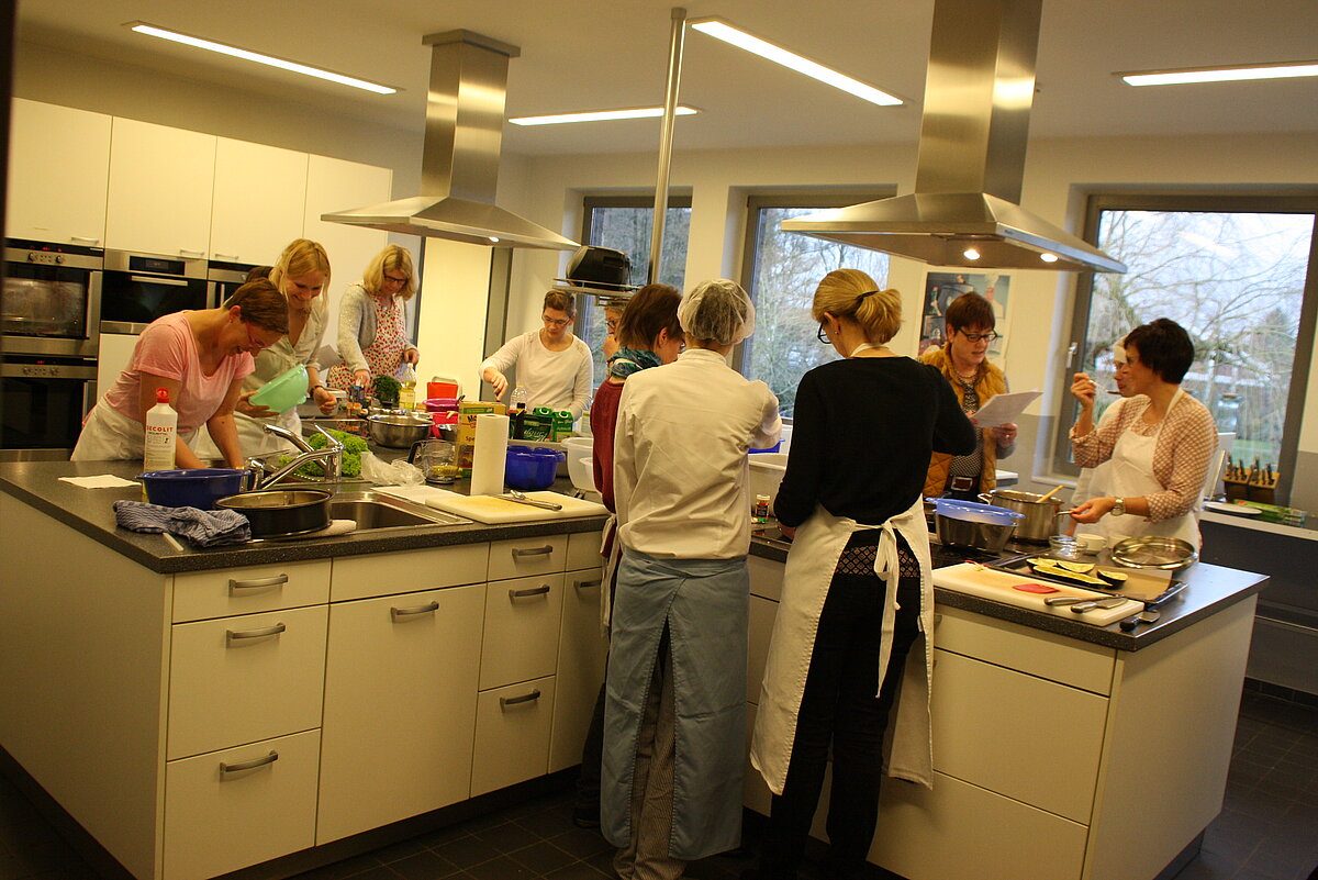 Küchenausstattung Ludgerus-Werk Eine große Kochinsel mit zwei Kochstellen. Drumherum steht eine Gruppe Kursteilnehmer, die kochen und Essen vorbereiten.