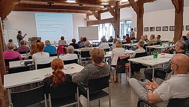 Die Hofstelle des Ludgerus-Werk. Die Teilnehmenden schauen nach vorne auf eine Leinwand, wo der Vortrag projiziert wird. Die Teilnehmenden sitzen an Tischen.  