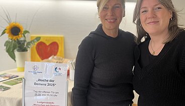 Marika Görtz-Ochotzki und Sabrina Diekhaus stehen an einem Stehtisch mit freundlicher Blumendekoration und Informationsblättern zu Menschen mit Demenz bewegen.