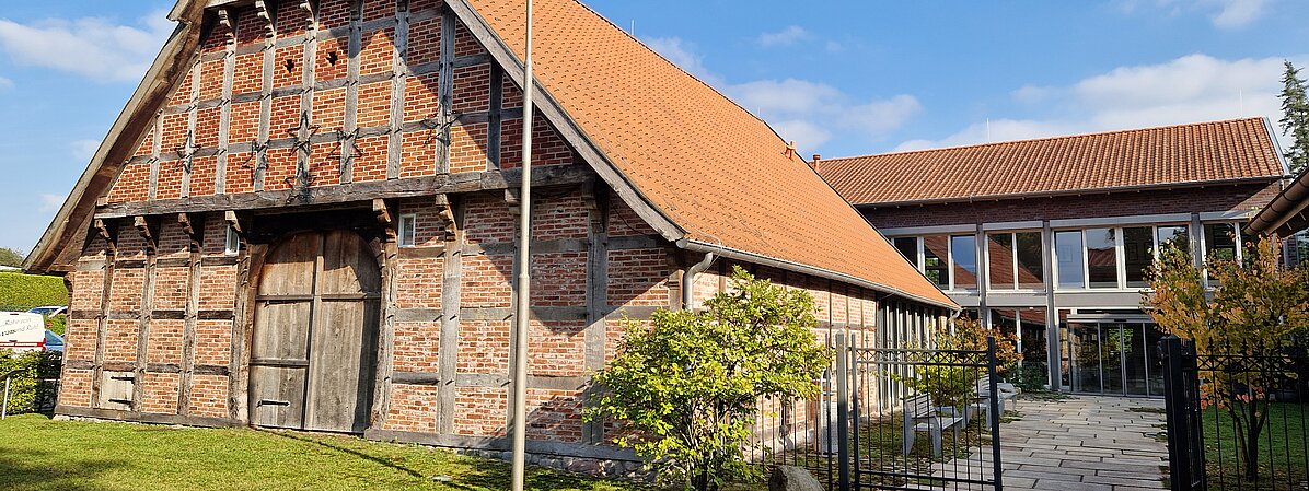 Header Ludgerus-Werk Hofstelle des Ludgerus-Werks, ein altes Fachwerkhaus auf der linken Seite, rechts ein metallener Eingangszaun, der auf das neugebaute Hauptgebäude zuführt
