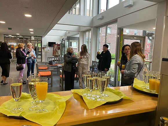 Im Vordergrund ist eine Theke zu sehen. Es stehen gefüllte Sektgläser darauf. Im Hintergrund stehen Besucher des Ludgerus-Werks. Sie unterhalten sich. 