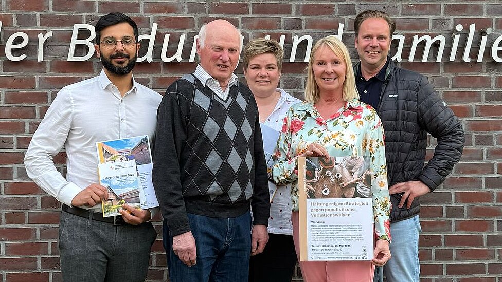 Eine Gruppe von 5 Leuten stehen vor dem Logo an der Außenwand des Ludgerus-Werk e.V. Lohne. Annette Kröger, die zweite Person von rechts, hält ein Plakat für den Vortrag "Haltung zeigen" von Marlies Stertenbrink in den Händen. 