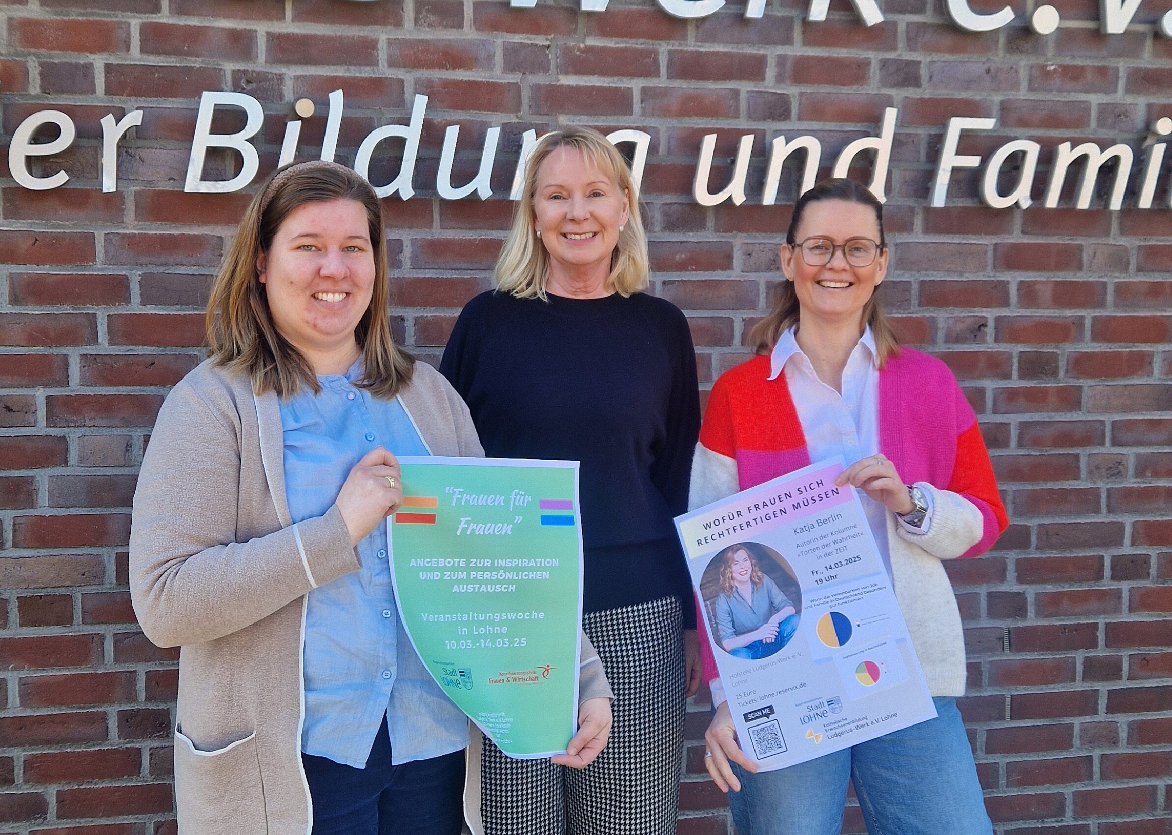 Drei lächelnde Frauen stehen vor dem Logo an der Außenwand des Ludgerus-Werk e.V. Lohne. V.l.n.r.: Rebecca Fischer hält ein Plakat für die Veranstaltungswoche für Frauen in den Händen, Annette Kröger in der Mitte, Nancy Menke hält ein Plakat von der Veranstaltung von Katja Berlin in den Händen.