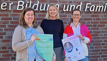 Drei lächelnde Frauen stehen vor dem Logo an der Außenwand des Ludgerus-Werk e.V. Lohne. V.l.n.r.: Rebecca Fischer hält ein Plakat für die Veranstaltungswoche für Frauen in den Händen, Annette Kröger in der Mitte, Nancy Menke hält ein Plakat von der Veranstaltung von Katja Berlin in den Händen.