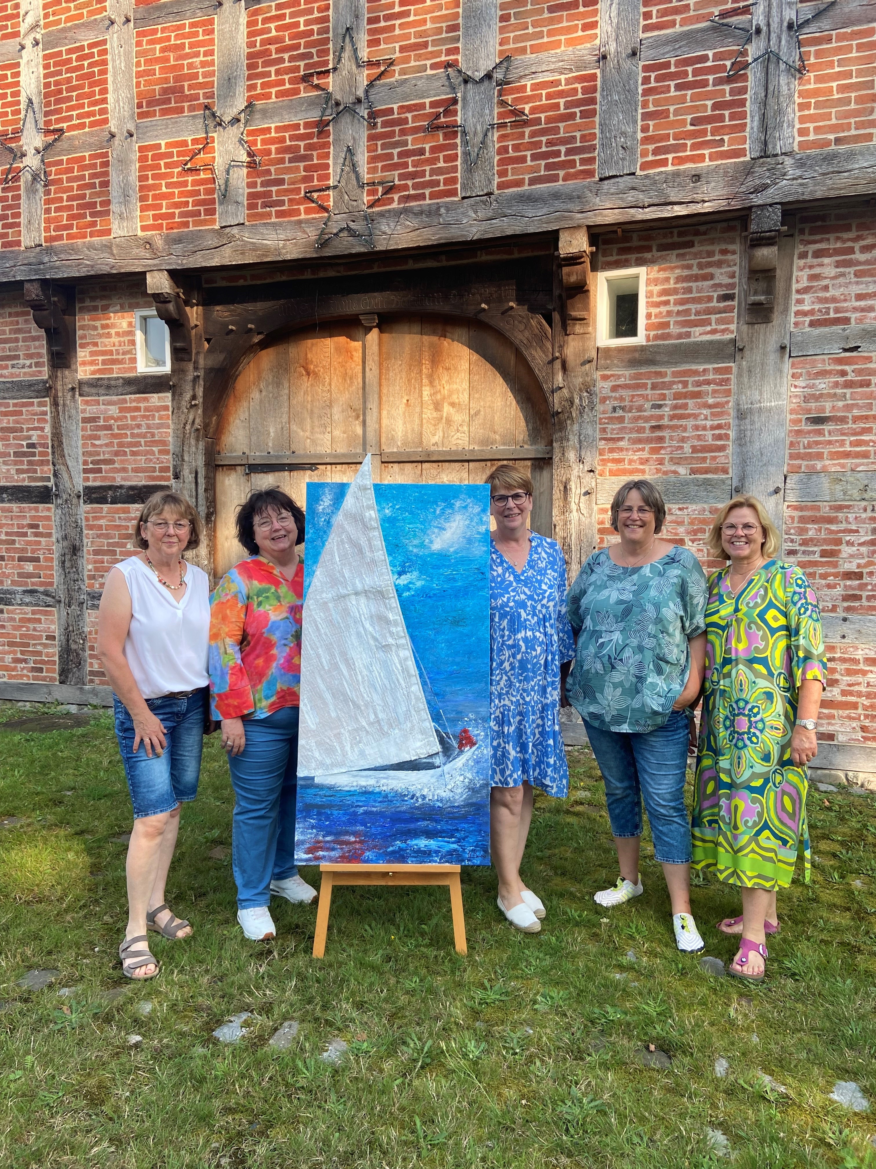 Fünf Künstlerinnen der Ausstellung Meer&Mehr stehen um ein Kunstwerk herum. Sie stehen draußen vor der Hofstelle des Ludgerus-Werks. Das Kunstwerk zeigt ein Segelboot auf dem Wasser. Das Segel des Bootes ist aus Stoff und ragt über den Rahmen hinaus. 