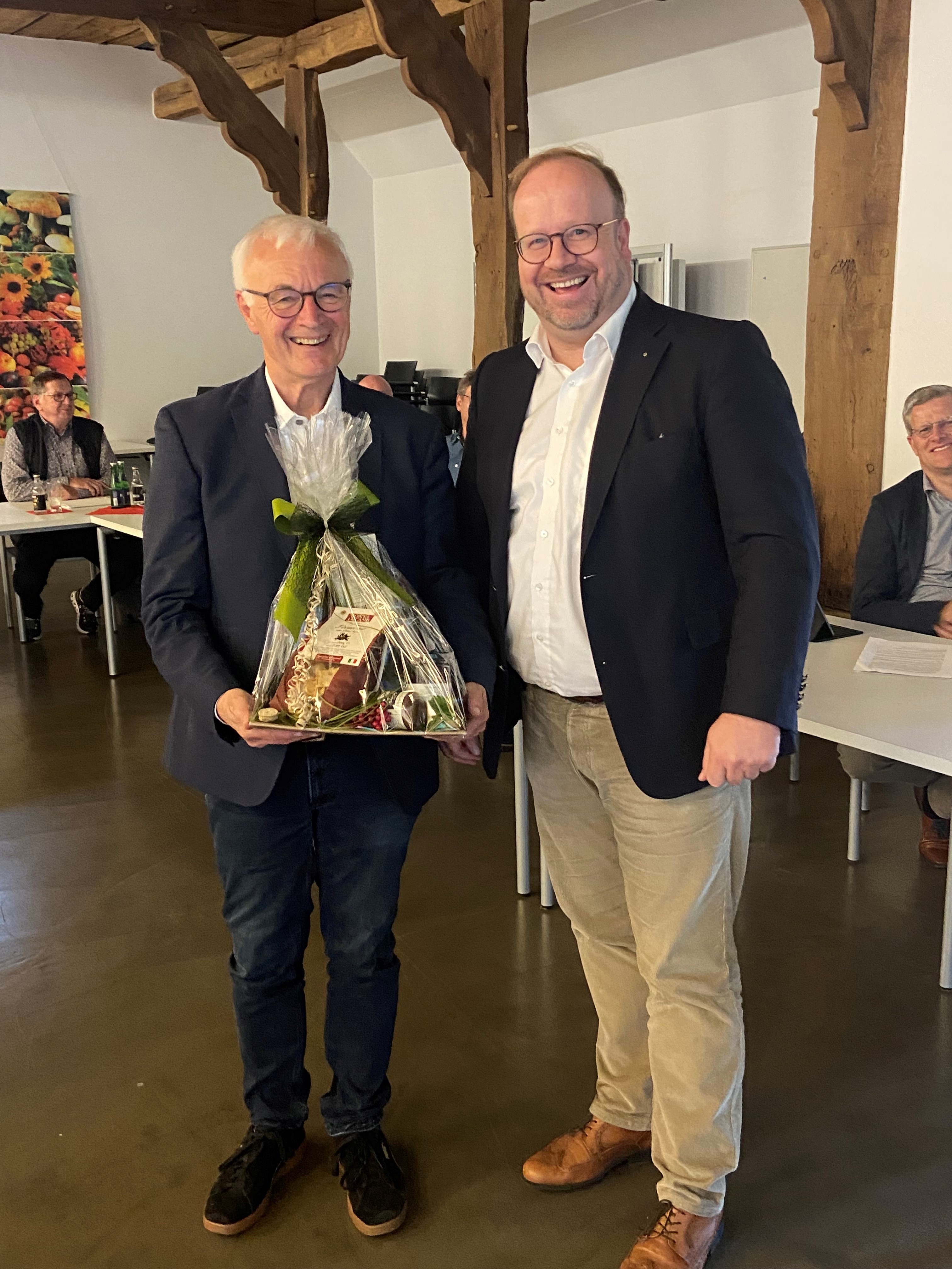 Zwei Männer stehen nebeneinander und lächeln in die Kamera. Der linke hält ein in Folie eingepacktes Geschenk in den Händen, das ihm überreicht worden ist. 
