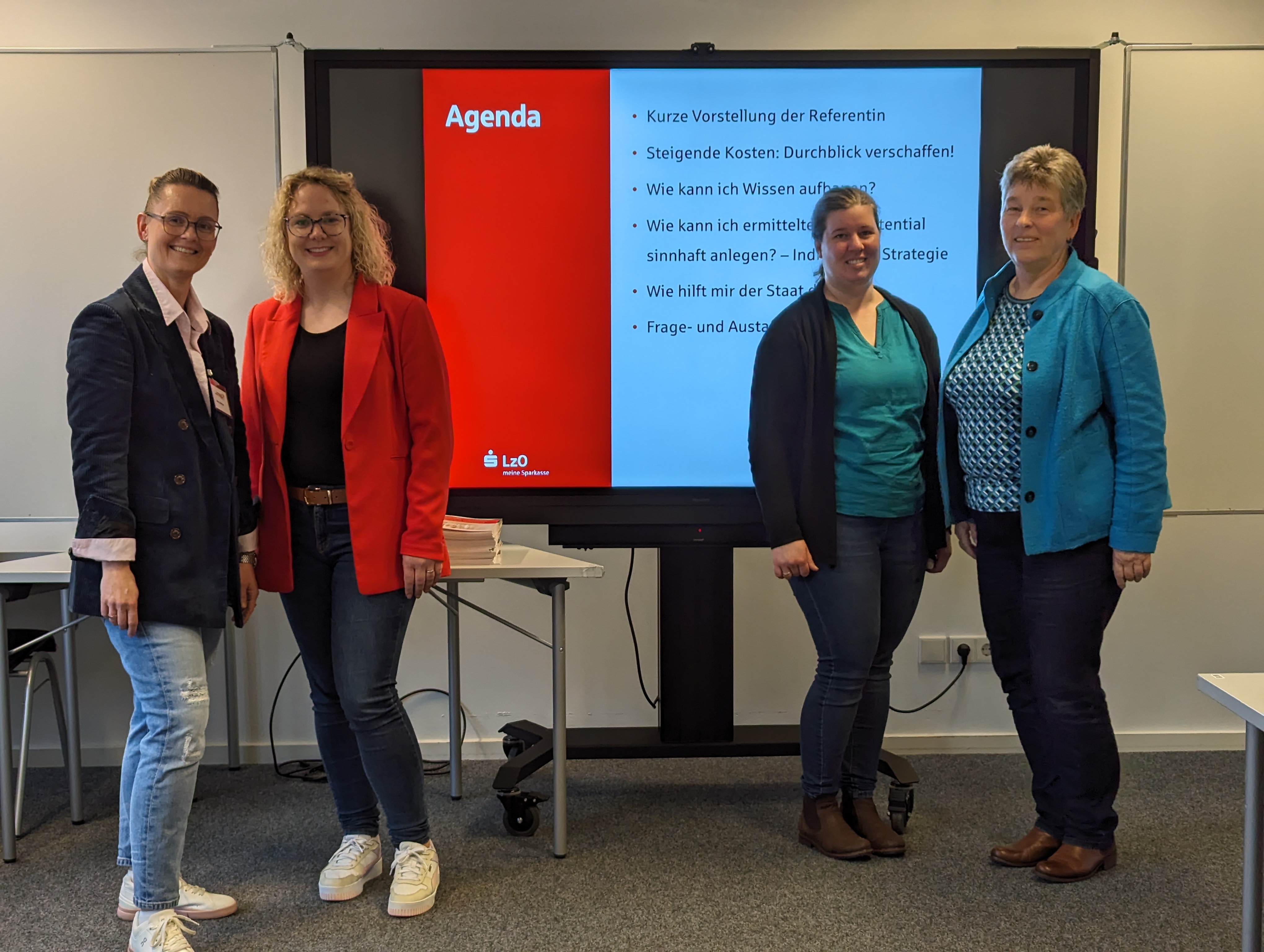 Vier Frauen stehen in jeweils in zwei Paaren neben einem Whiteboard. Sie schauen lächelnd in die Kamera. Auf dem Whiteboard ist die Agenda des Vortrags abgebildet.