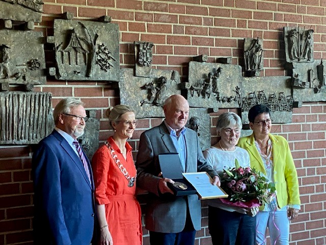 Eine Gruppe Leute stehen um den geehrten Werner Becker vor einer Ziegelwand mit Bildern daran. Werner Becker steht in der Mitte und hält eine Urkunde und eine Medaille in einem Kästchen in der Hand.