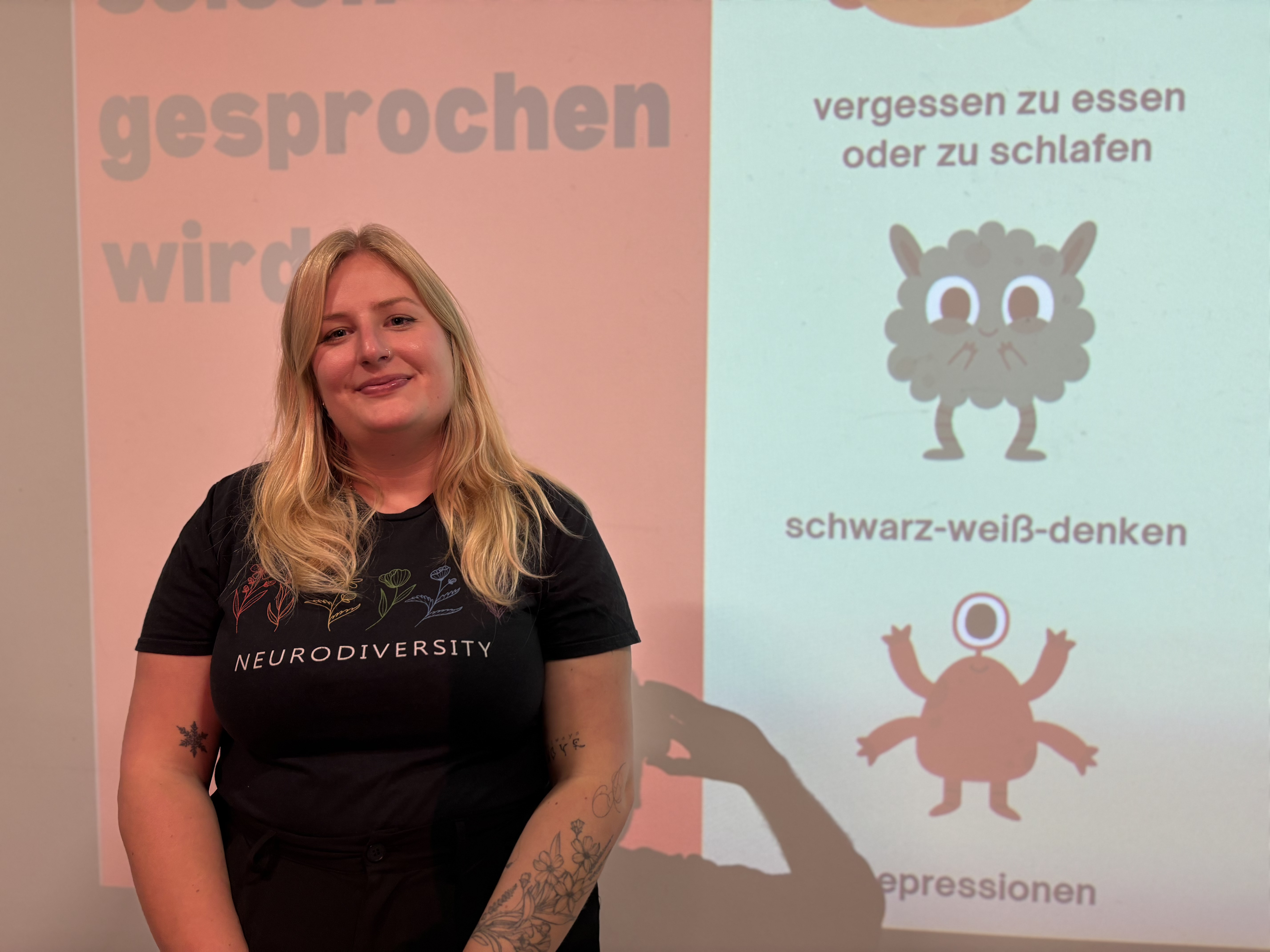 Eine blonde Frau steht vor einer Leinwand. Auf die Leinwand sind Teile ihres Vortrags über Autismus und ADHS projiziert. 