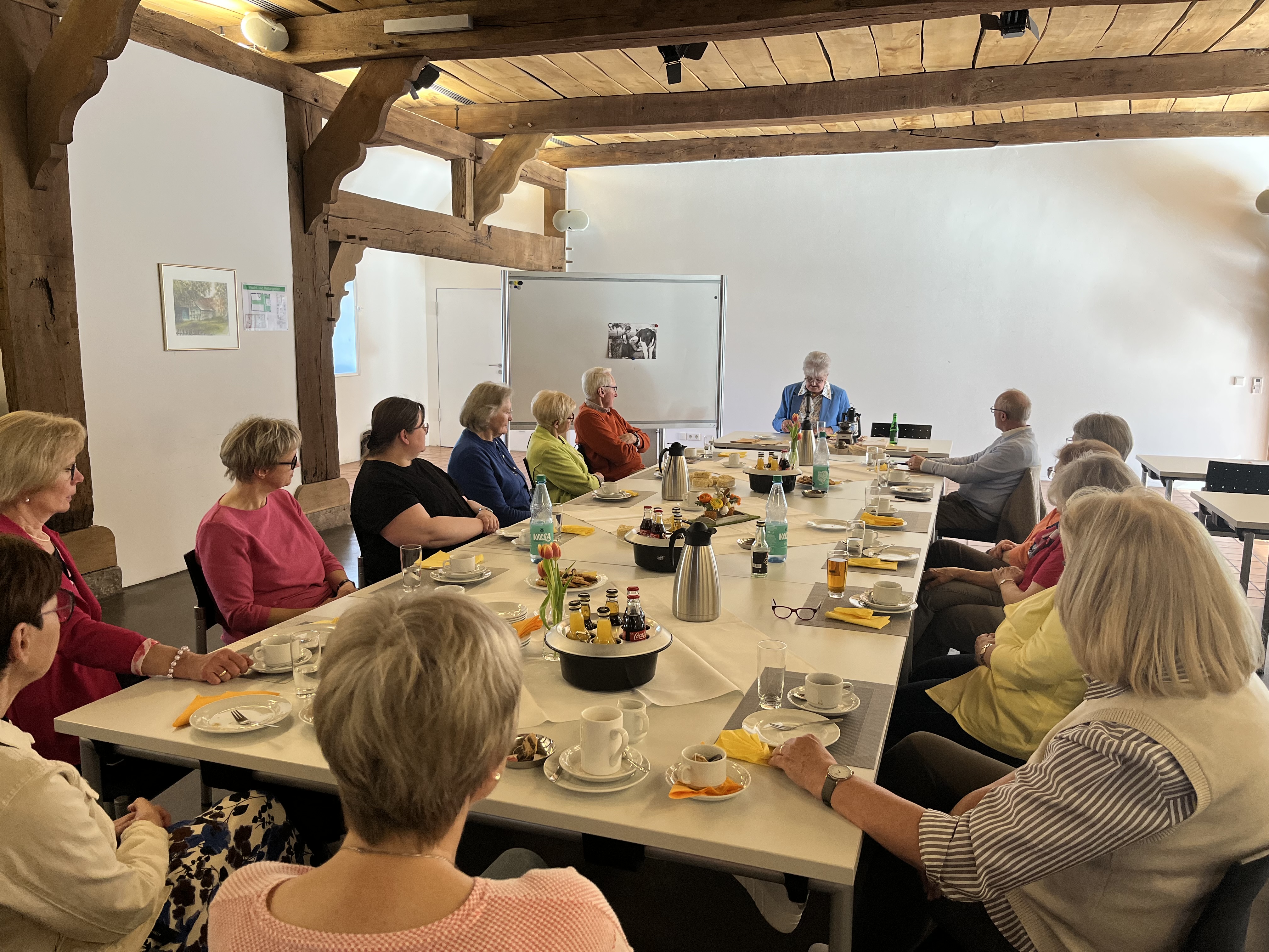 Ein großer Tisch in der Hofstelle im Ludgerus-Werk an dem alle in der Runde sitzen. Es ist für Kaffee und Kuchen gedeckt. An der Stirnseite sitzt die Autorin Maria Meyer, die aus ihrem Buch vorliest.