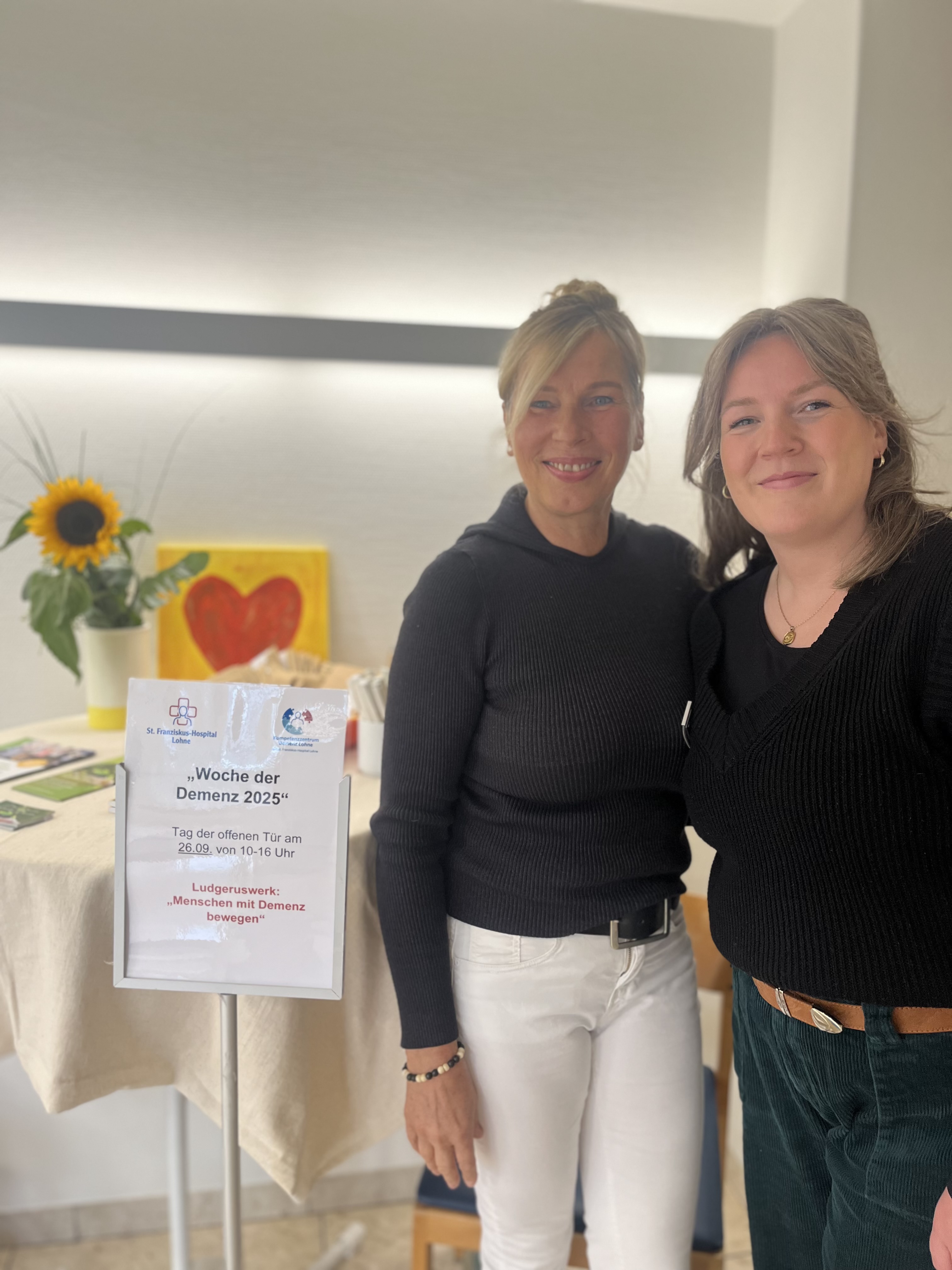 Marika Görtz-Ochotzki und Sabrina Diekhaus stehen an einem Stehtisch mit freundlicher Blumendekoration und Informationsblättern zu Menschen mit Demenz bewegen.