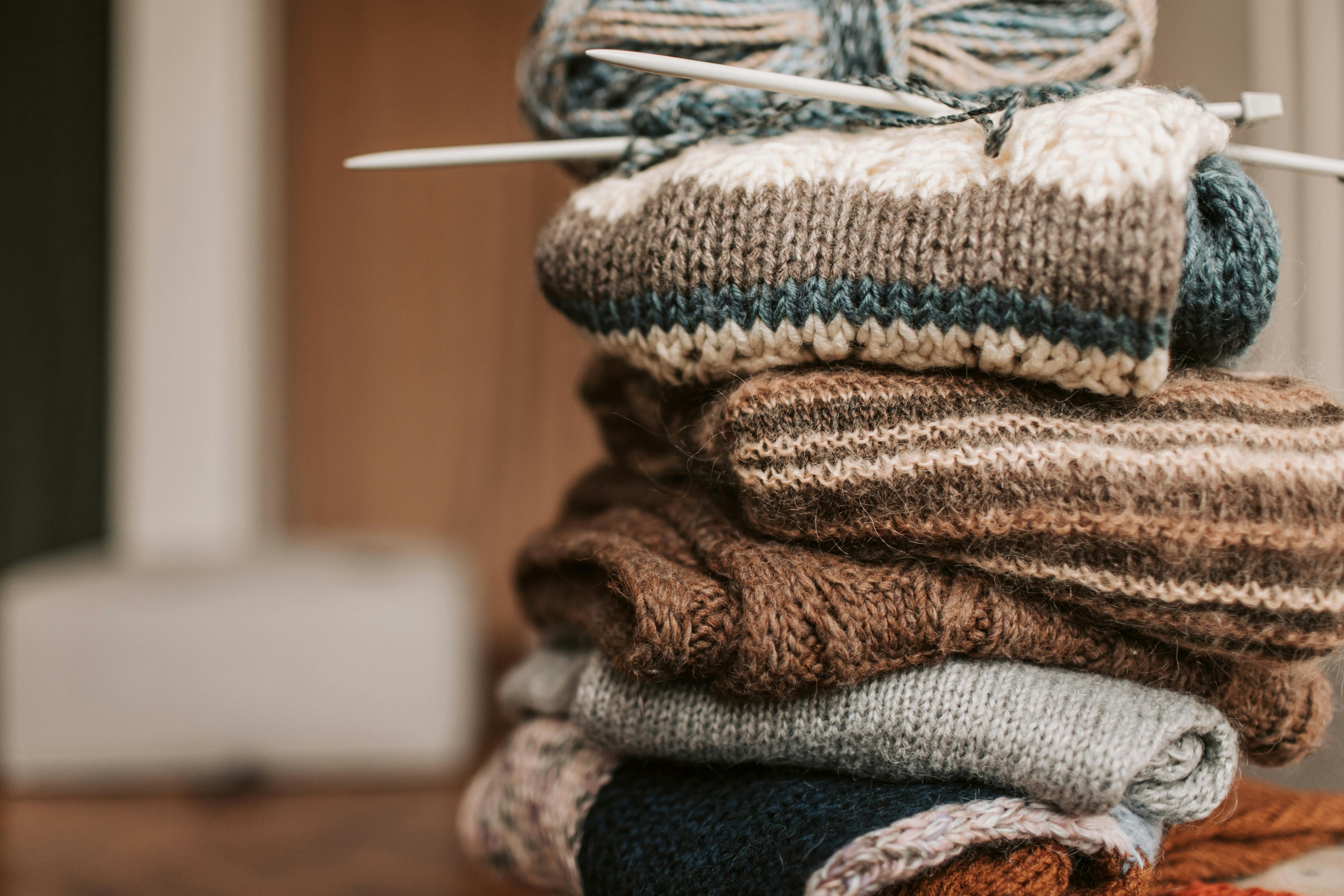 Ein Stapel gestrickter Objekte die zusammengelegt auf einem Tisch liegen. Obenauf liegt ein Knäuel Wolle mit Stricknadeln. 